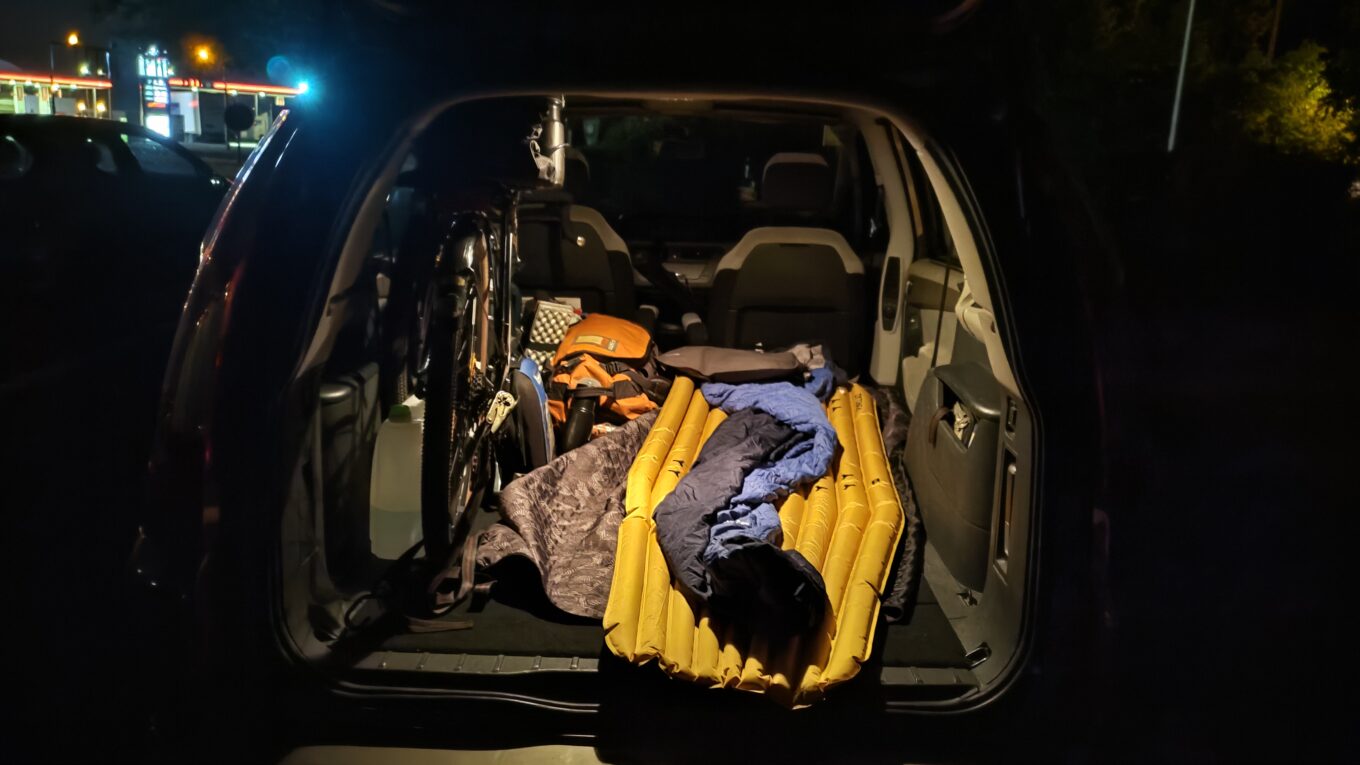 Vue de l'arrière d'un hayon de voiture de nuit. Dans le coffre, sièges arrières rabattus, un vélo et à côté un matelas gonflable et un duvet