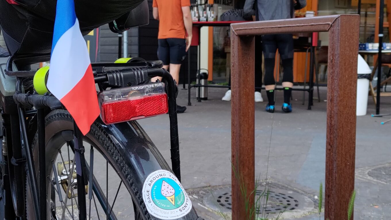 Vue verticale de l'arrière d'un vélo équipé en bikepacking mais aussi un garde boue et un porte bagages. Sur le garde-boue plusieurs autocollants dont #ratngx @BiclouneParis, sur le porte bagages un petit drapeau Français. En arrière plan un abris en toile au logo de De Proloog