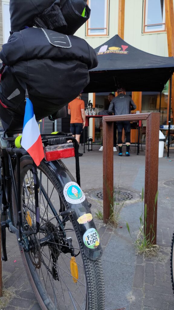 Vue verticale de l'arrière d'un vélo équipé en bikepacking mais aussi un garde boue et un porte bagages. Sur le garde-boue plusieurs autocollants dont #ratngx @BiclouneParis, sur le porte bagages un petit drapeau Français. En arrière plan un abris en toile au logo de De Proloog