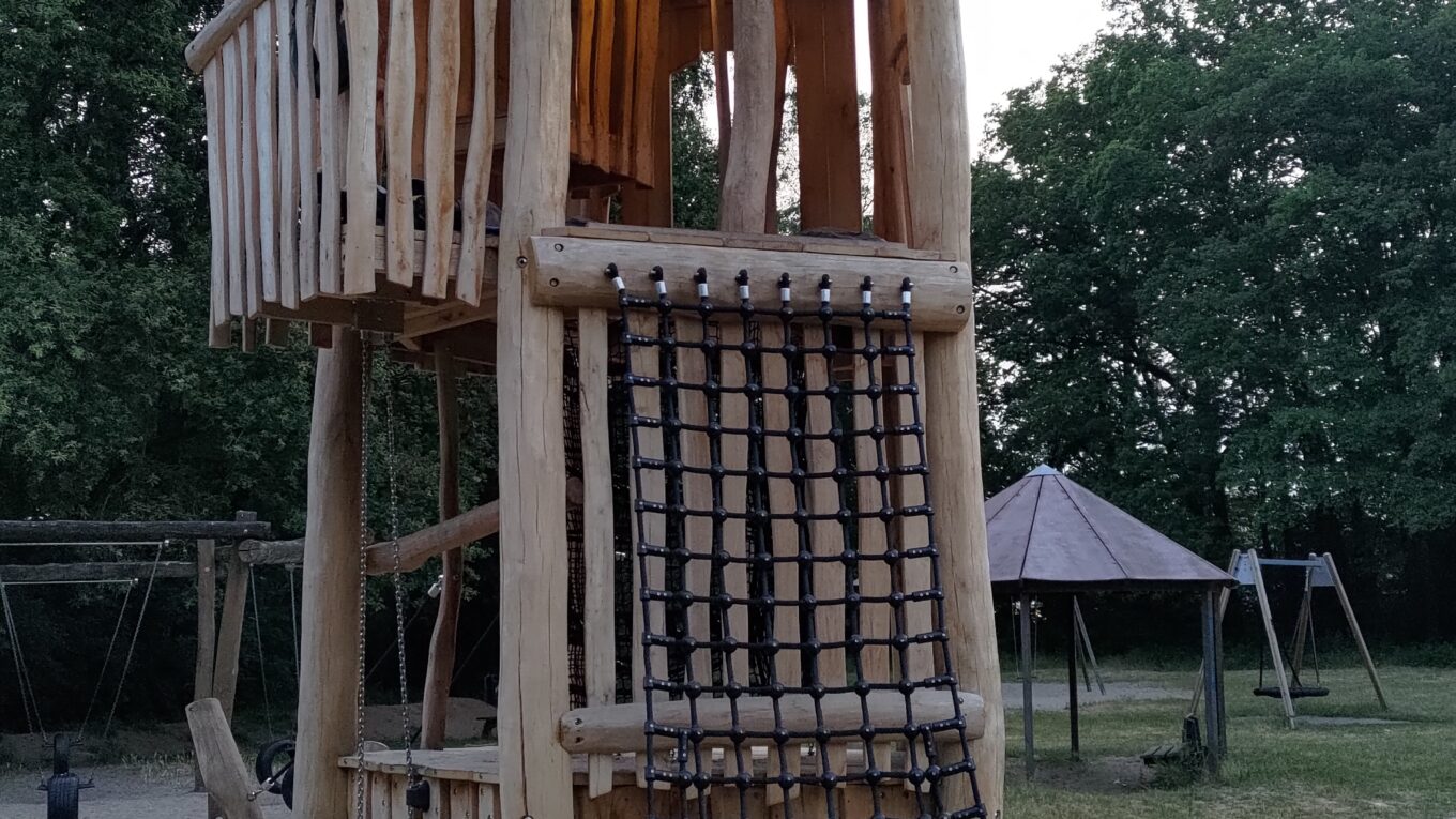 Tour de 4 étages en bois au milieu d'un bac à sable dans un parc pour enfant. Au pied de la tour un vélo attaché à une échelle de cordes. Au 3ème étage une lampe éclaire le plafond