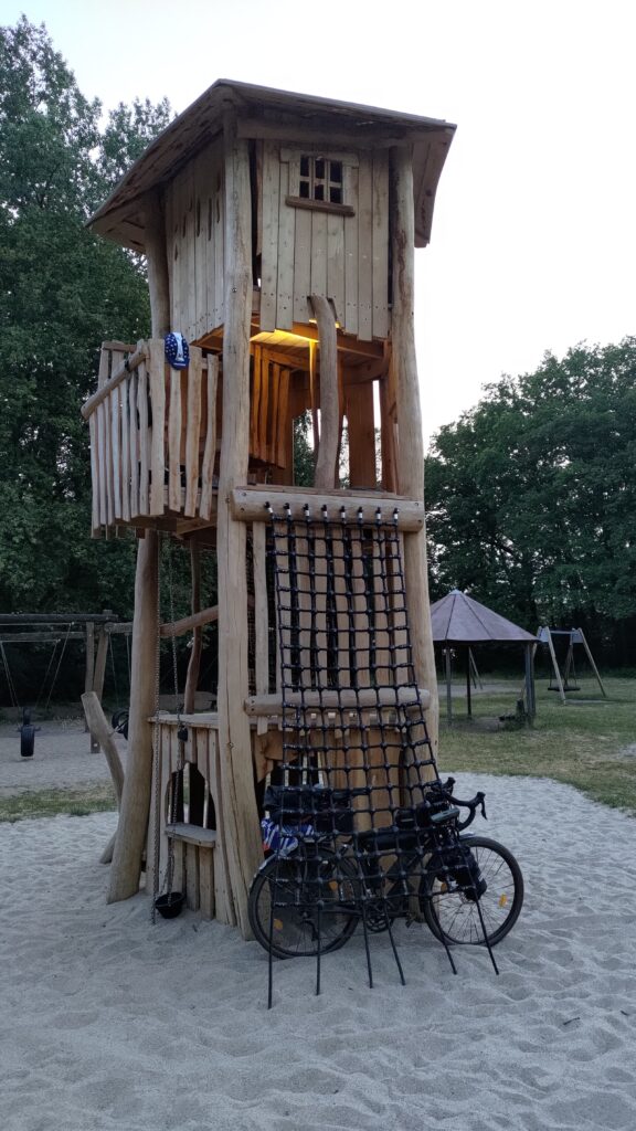 Tour de 4 étages en bois au milieu d'un bac à sable dans un parc pour enfant. Au pied de la tour un vélo attaché à une échelle de cordes. Au 3ème étage une lampe éclaire le plafond