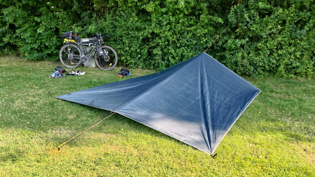 Au premier plan un abris en forme de tente fabriqué avec un tarp, en arrière plan un vélo le long du haute haie