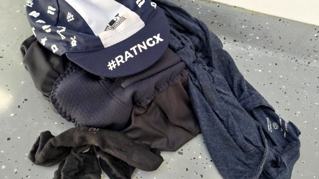 Des vêtements manifestement mouillés posés sur le sol d'une douche. On distingue entre autres un cuissard de cycliste. Posé sur le dessus une casquette bleue et blanche siglée #ratngx