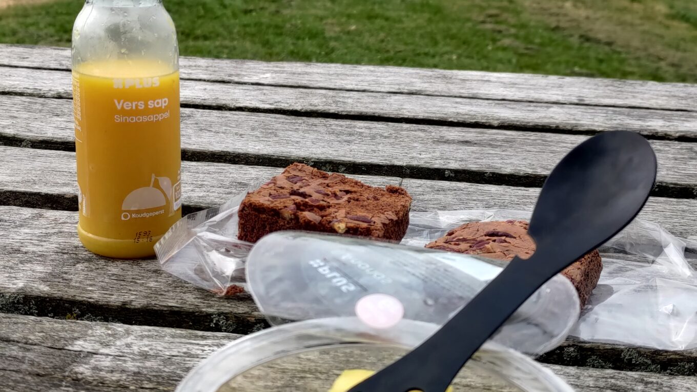 Posés sur une table de picnic, une boîte d'ananas en morceaux, une bouteille de jus d'orange et deux parts de brownie. En arrière plan un moulin à vent