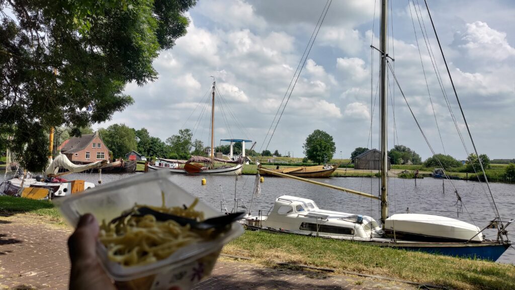 Petit port sur un canal et une barquette de pâtes tenue à une main