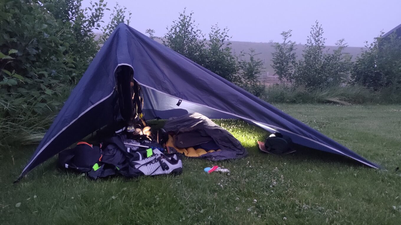 Tente improvisée. Un vélo sert de support, par-dessus un tarp permet de créer un abri. Sous le tarp, un.i. vy,un matelas et tout le reste du matériel