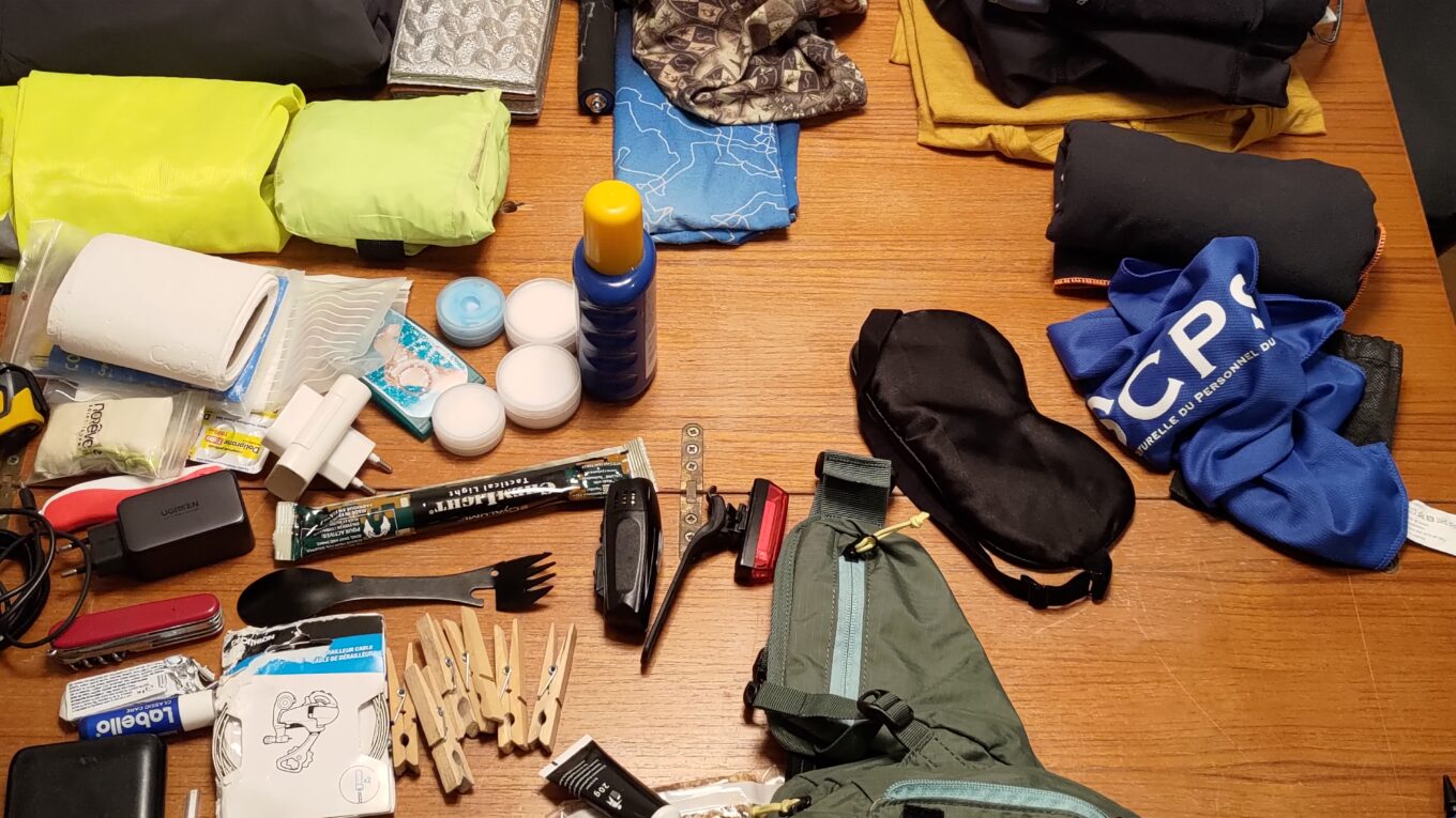 Plan serrée sur une table en bois vue du dessus. De très nombreuses affaires sont disposées sur toute la surface : vêtements, bidons, casque, matériel de couchage, batteries, câbles, lampes, barres de céréales, nécessaire de toilette, etc…