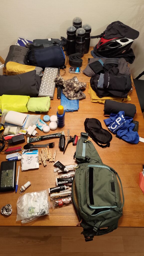 Plan serrée sur une table en bois vue du dessus. De très nombreuses affaires sont disposées sur toute la surface : vêtements, bidons, casque, matériel de couchage, batteries, câbles, lampes, barres de céréales, nécessaire de toilette, etc…