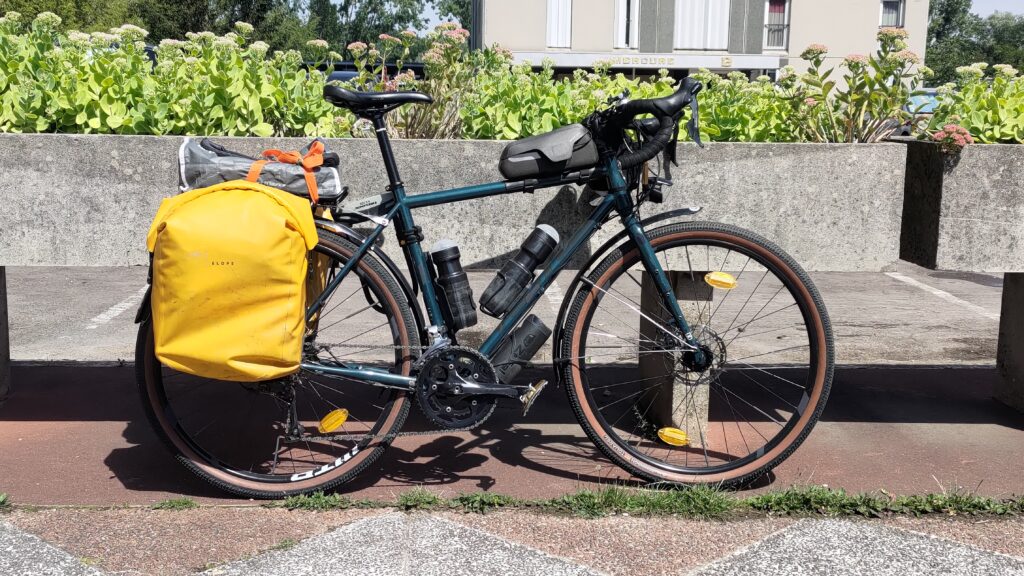 Velo type gravel équipé avec des sacoches jaunes, manifestement pleines et un paquet sur le porte bagages. Bidons et food pouch.