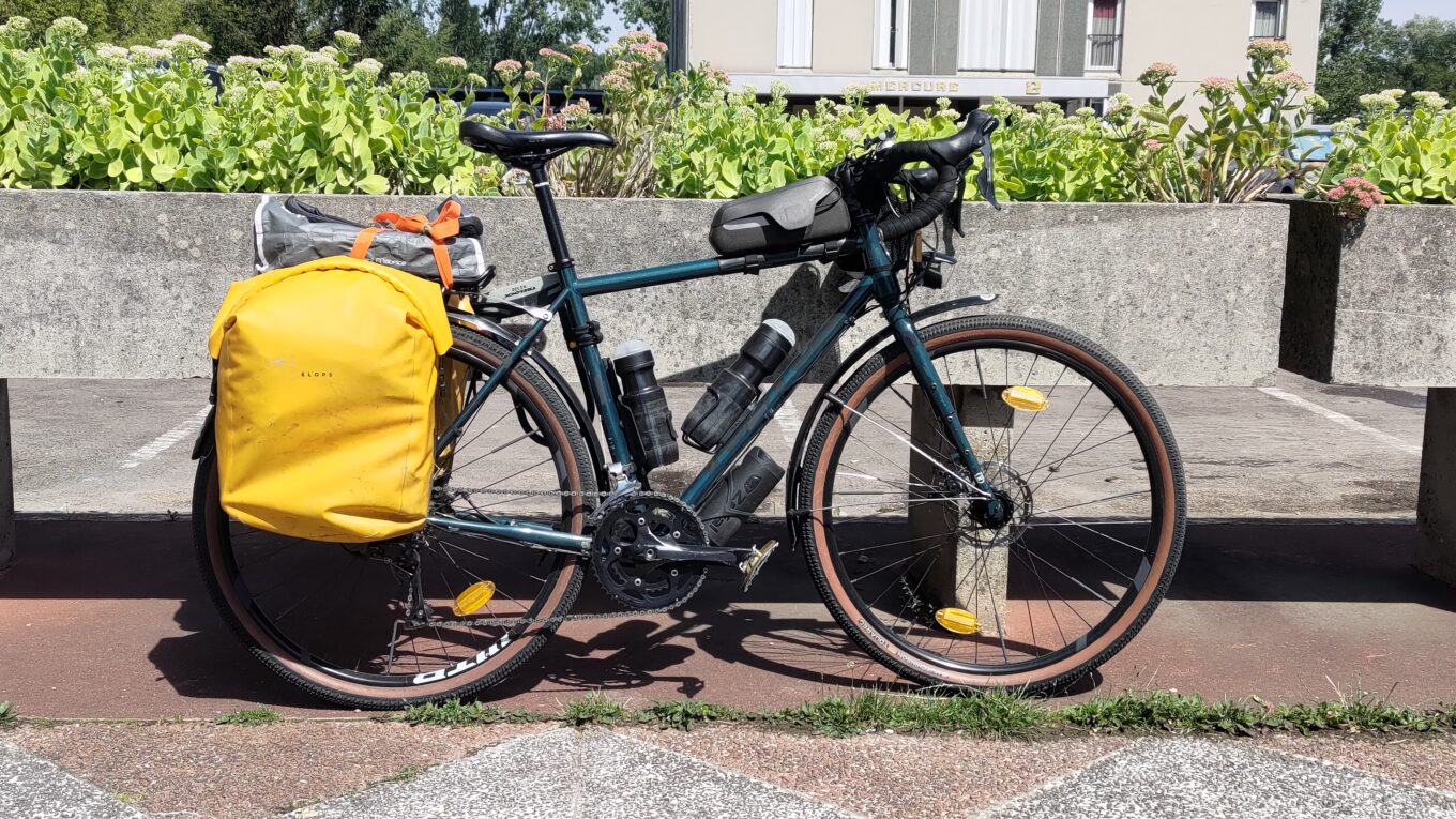 Velo type gravel équipé avec des sacoches jaunes, manifestement pleines et un paquet sur le porte bagages. Bidons et food pouch.