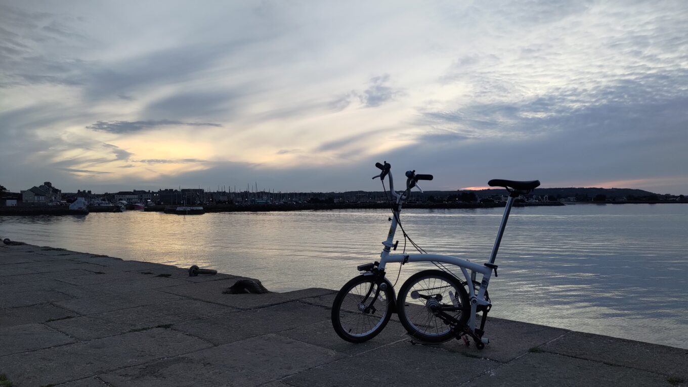 Velo pliant à moitié plié posé sur une dingue. Au premier plan derrière le vélo la mer, derrière le port et la ville. Le soleil se couche et le ciel chargé de quelques nuages à de belles couleurs jaune gris bleu et un peu de rouge