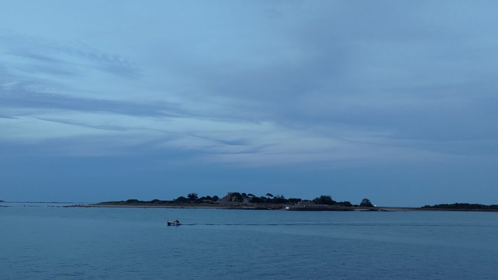 Dans la pénombre une île au loin occupe tout l'horizon. Un petit bateau passe