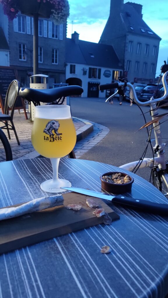 Table de bar en extérieur avec un verre de bière, un saucisson sur une planche à découper er des cacahuètes. Derrière la table un vélo pliant. En arrière plan des maisons. Pénombre.