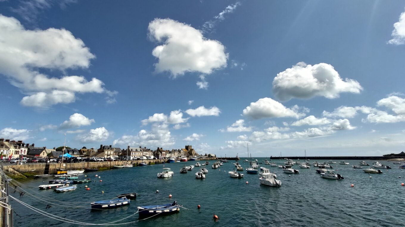 Vue sur le port de Barfleur. Plusieurs bateaux de plaisance ou de pêche sont amarrés sur le plan d'eau, des maisons sur le quai visibles sur la gauche. Beau ciel bleu et gros nuages blancs