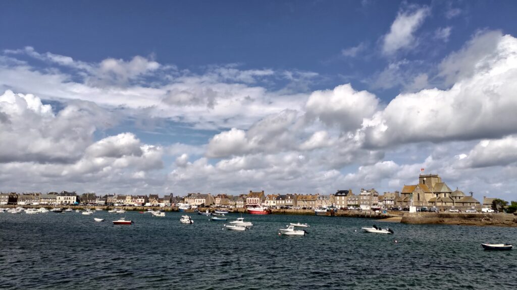 Vue sur le port de Barfleur. Plusieurs bateaux de plaisance ou de pêche sont amarrés sur le plan d'eau, des maisons sur le quai visibles sur la gauche. Beau ciel bleu et gros nuages blancs