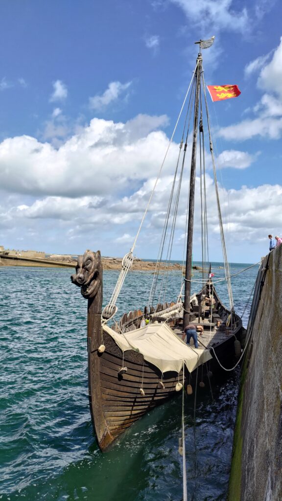 Un drakkar en bois amarré sur un quai, un drapeau normand flotte en haut du mat.