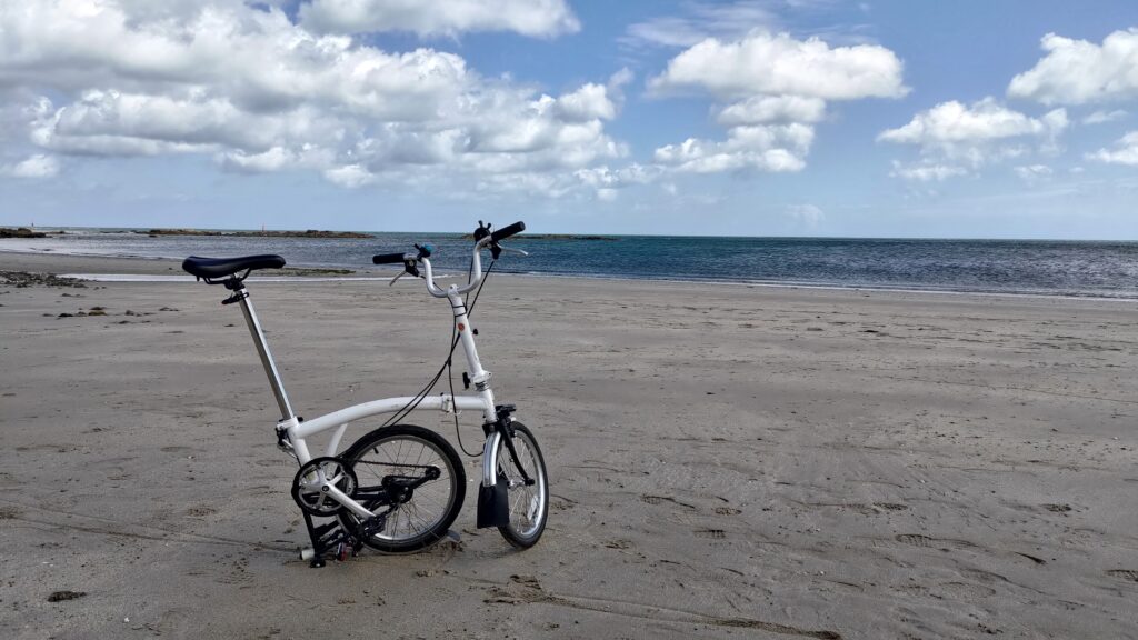 Un Brompton à moitié plié sur une plage de sable, la mer au loin