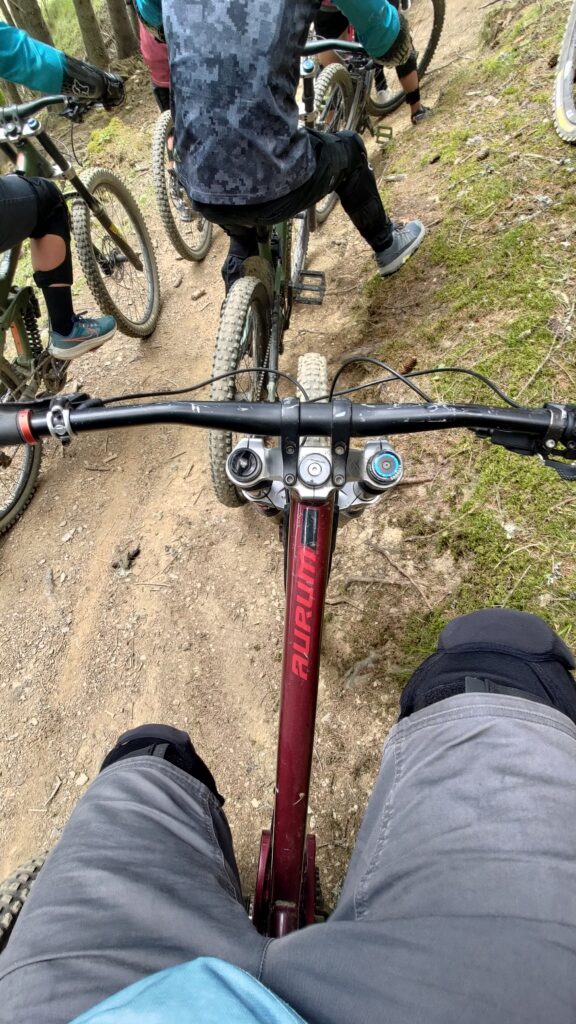 Vue subjective depuis un cycliste assis sur un VTT. On voit ses cuisses et le cintre du vélo et le sol en terre. Les jambes et vélos d'autres cyclistes sont également visibles