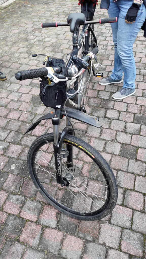 Un vélo tandem VTT vu de devant. On voit la fourche suspendue. Personne n'est assis sur la selle avant et le vélo est maintenue par la selle arrière par une personne dont on ne voit que les jambes habillées d'un jean. Des pavés roses et sales au sol.