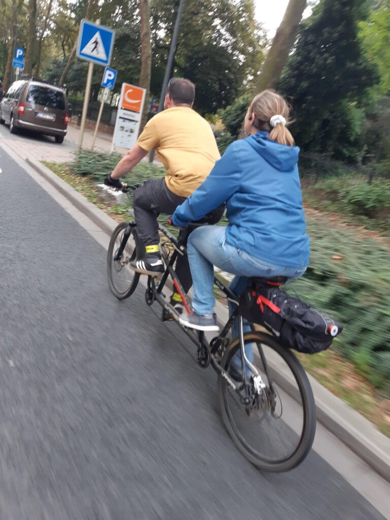 Deux personnes pédalent sur un tandem VTT dans un environnement urbain mais boisé