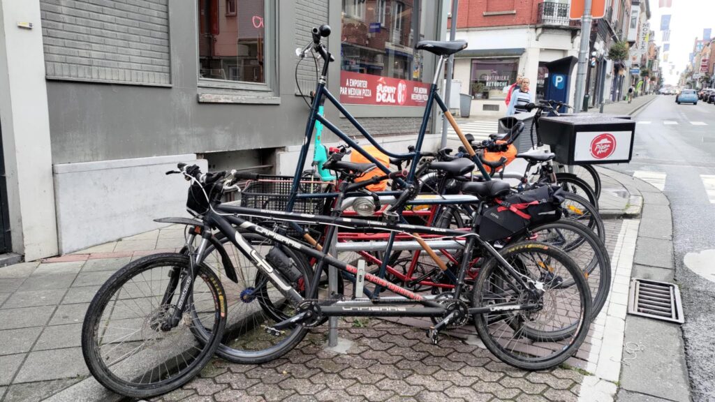 Vu d'un parking vélos le long d'un trottoir. Il est difficile de distinguer tous les vélos (une dizaine) mais au premier plan un tandem, ensuite un tallbike, ensuite un vélo cargo Petit Porteur. Le reste est plus confus.