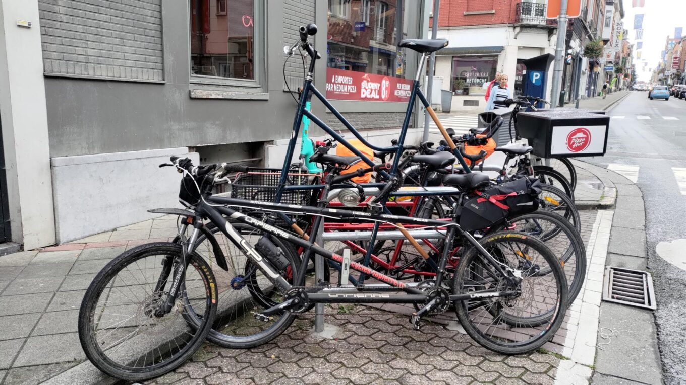 Vu d'un parking vélos le long d'un trottoir. Il est difficile de distinguer tous les vélos (une dizaine) mais au premier plan un tandem, ensuite un tallbike, ensuite un vélo cargo Petit Porteur. Le reste est plus confus.