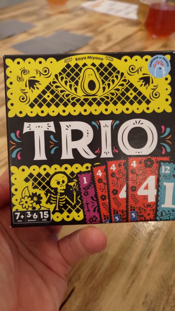 Photo d'une boite de jeu de société tenue dans une main. Le jeu est Trio par Kaya Miyano, 7+ ans, 3-6 joueurs, 15 min. La boite est décorée de façon vive avec un thème rappelant le Día de Muertos mexicain, des squelettes et des motifs végétaux stylisés.