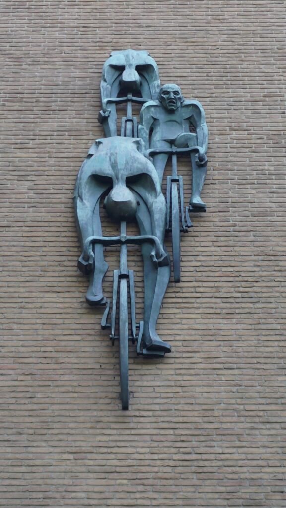 Sculpture en bronze murale sur un mur de briques de couleur crème. Trois cyclistes stylisés sur leur vélo, vu de face. Deux baissent la tête en position de recherche de vitesse, le troisième à le visage grimaçant d'effort. Cette sculpture est sur le mur du Vélodrome Kuipke de Gand