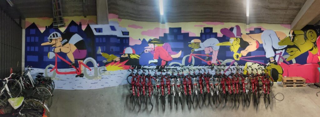 Fresque mural très colorée représentant plusieurs cyclistes les uns derrières les autres dans un style bande dessinée. Dans un sous-sol parking. Plusieurs vélos du même modèle d'une entreprise de location sont garés alignés perpendiculairement au mur.