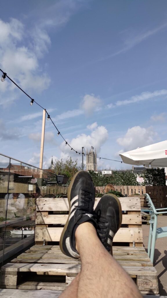 Au premier plan le bas des jambes nues et poilues, croisées et des chaussures noires, en second plan un banc en palette et dans l'axe des jambes une tour médiévale.