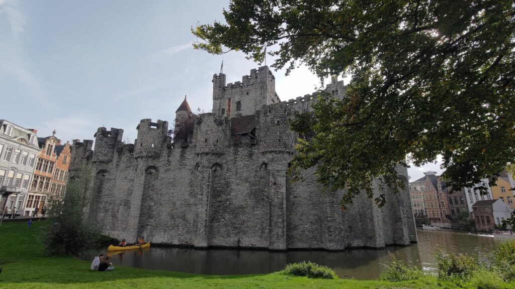 Vu d'un château médiéval avec des douves, un kayak jaune est dans l'eau. Les murs sont hauts et gris, le haut d'un donjon est visible dans l'enceinte fortifiée. Ce château est en pleine ville, on voit à droite et à gauche des maisons de style hollandais. Au premier plan de l'herbe d'un parc et sur la droite un grand arbre bouche la vue.