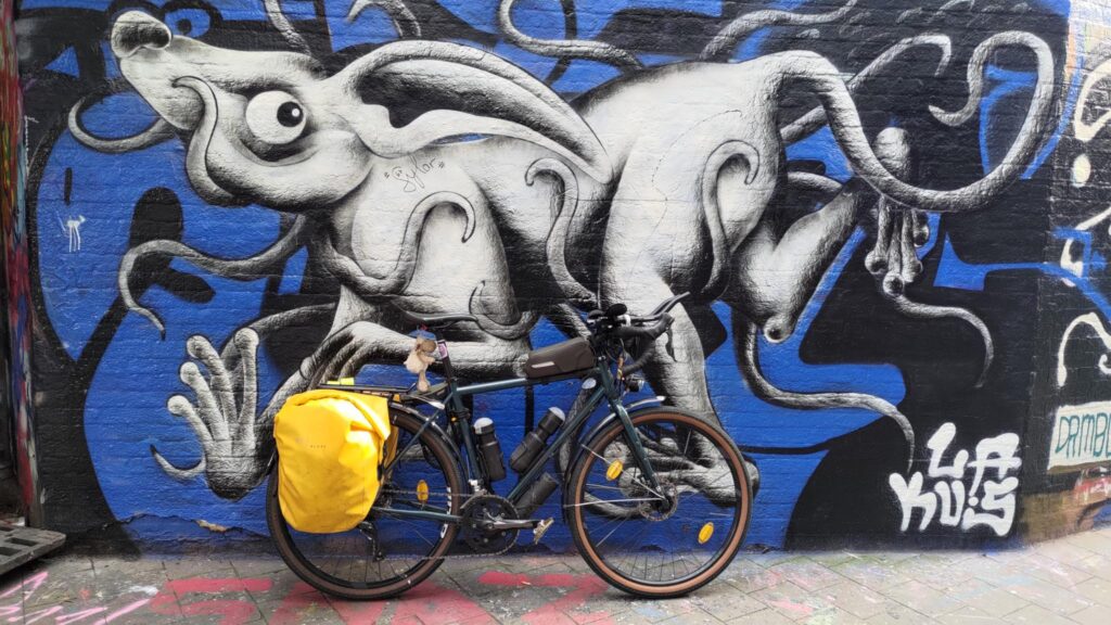 très grande fresque murale sur fond bleu et noir. Un animal imaginaire inspiré d'un lapin et d'un chien avec des tentacules est dessiné en blanc et ombrage gris et occupe toute la surface du mur. Un vélo type gravel est posé sur le mur avec des sacoches arrières jaunes.