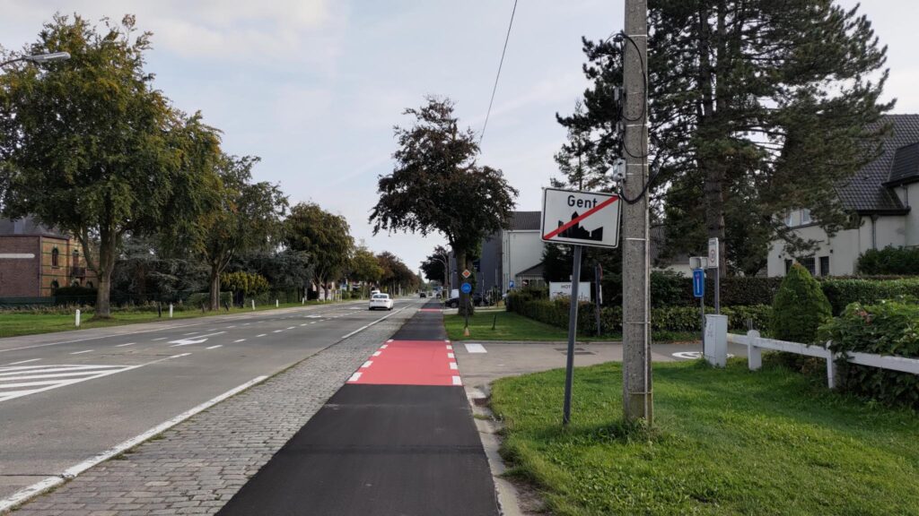 Vue en perspective d'une piste cyclable toute droite. Sur la gauche de la piste des pavés et la route, sur la droite de l'herbe et une intersection. Un panneau de sortie de ville barrée indique Gent. La piste cyclable a un revêtement en bitume noir neuf et les intersections sont rouges très visibles.