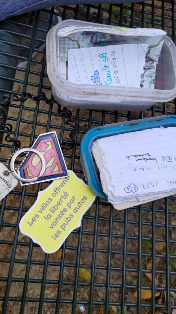 Posés sur une grille, le contenu d'une boite en plastique ouverte avec notamment un petit carnet de feuilles avec des inscriptions, un porte clé reprenant le logo de Spideman et autocollant jaune et texte violet "Les vélos offrent la liberté vantée par les pubs autos". C'est une géocache.