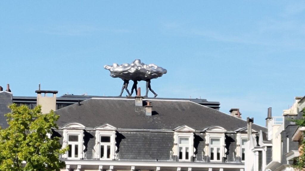 Toit d'immeuble du début du 20ème siècle avec dessus une sculpture moderne représentant un nuage métallique gris qui a trois paires de jambes.