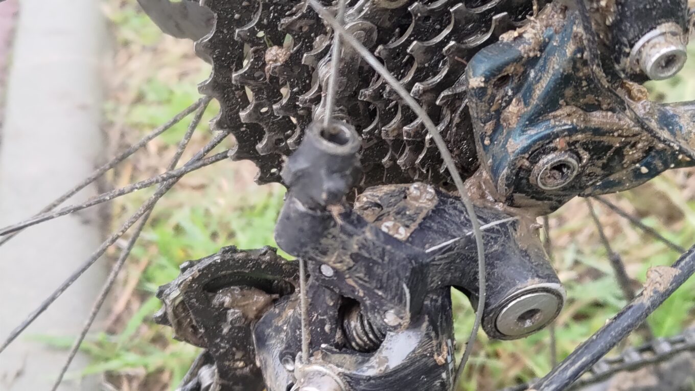 Plan serré sur le dérailleur d'un vélo. Le câble du dérailleur est entouré sur le cadre au lieu de passer dans la gaine. C'est un bricolage de fortune.