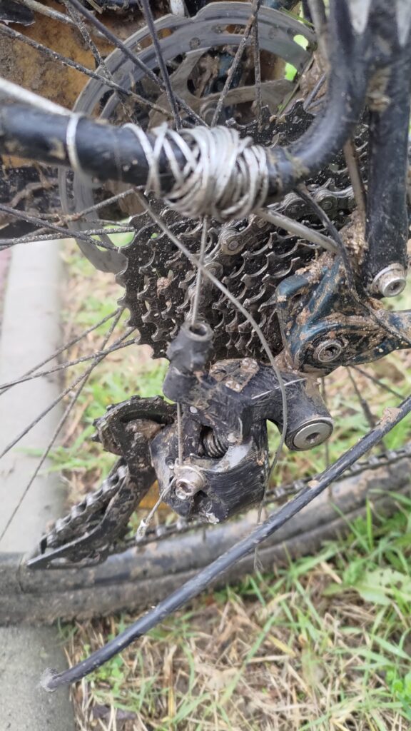 Plan serré sur le dérailleur d'un vélo. Le câble du dérailleur est entouré sur le cadre au lieu de passer dans la gaine. C'est un bricolage de fortune.