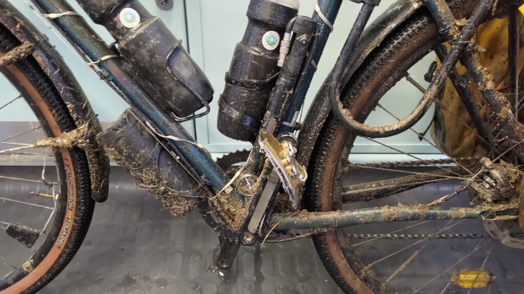 Plan serré sur le centre d'un vélo. On voit partiellement les roues, des bidons, un U et surtout beaucoup de boue