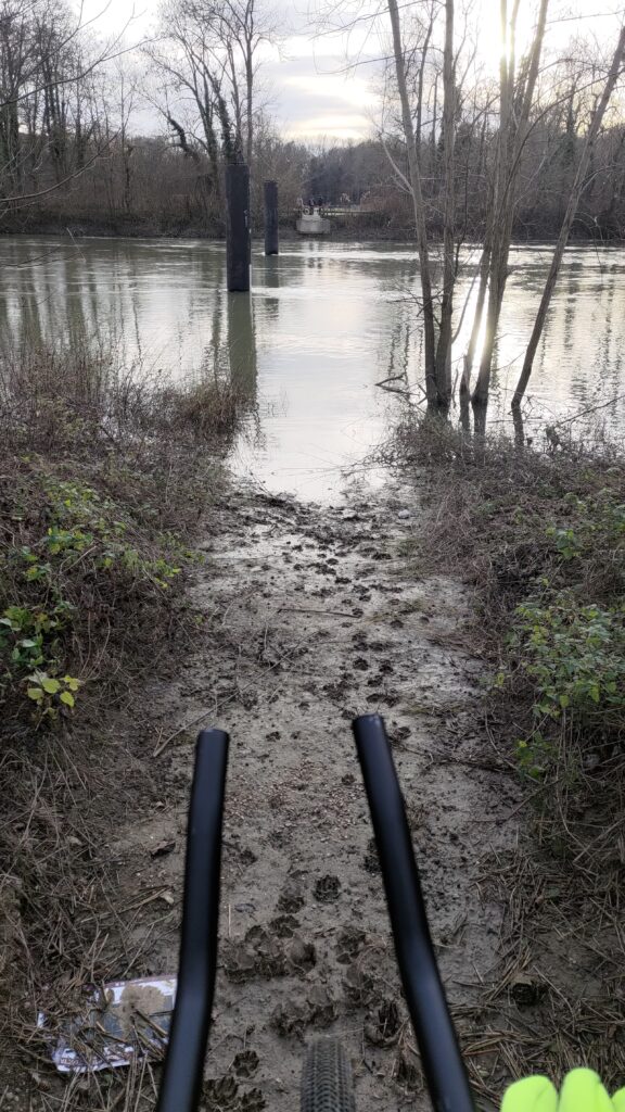L'avant d'un vélo est visible devant une rivière (la Marne) et deux pylônes sont visibles dans l'eau, montrant la présence habituelle d'un pont. D'autres cyclistes regardent également sur l'autre rive.
