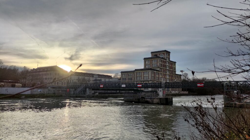 Vue sur la Marne, un pont métallique traverse la rivière. À l'arrière plan un grand bâtiment industriel sur trois niveaux mais chaque niveau est plus étroit que celui d'en dessous. D'autres bâtiments autour font parti du site industriel (ancienne usine de chocolat Menier à Noisiel)