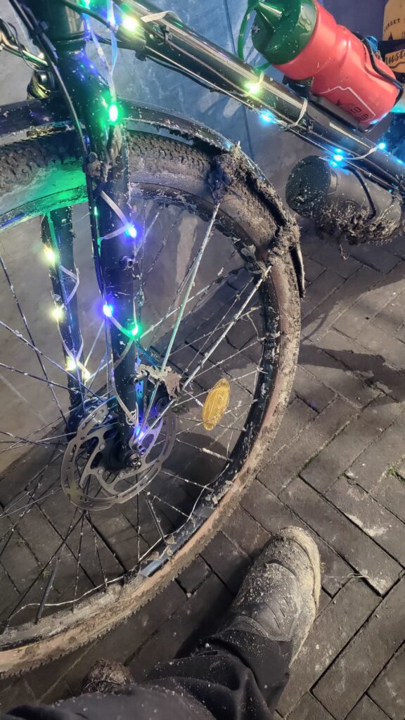 De nuit, plan sur l'avant d'un vélo type gravel couvert de boue et un pied du cycliste est également couvert de boue. Accessoirement le vélo est décoré de guirlandes de Noël et de deux gourdes Ketchup (rouge) et Moutarde (jaune)