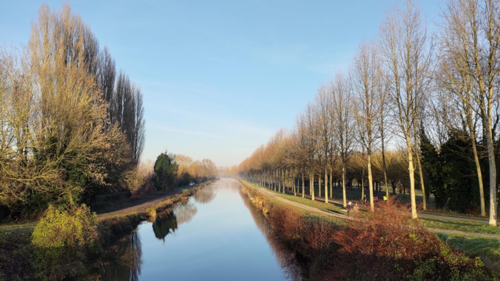 Vue sur la Somme depuis un pont, dans la perspective de la rivière. Grand ciel bleu. Sur la droite une rangée de hauts arbres avec un chemin et des piétons et des cyclistes