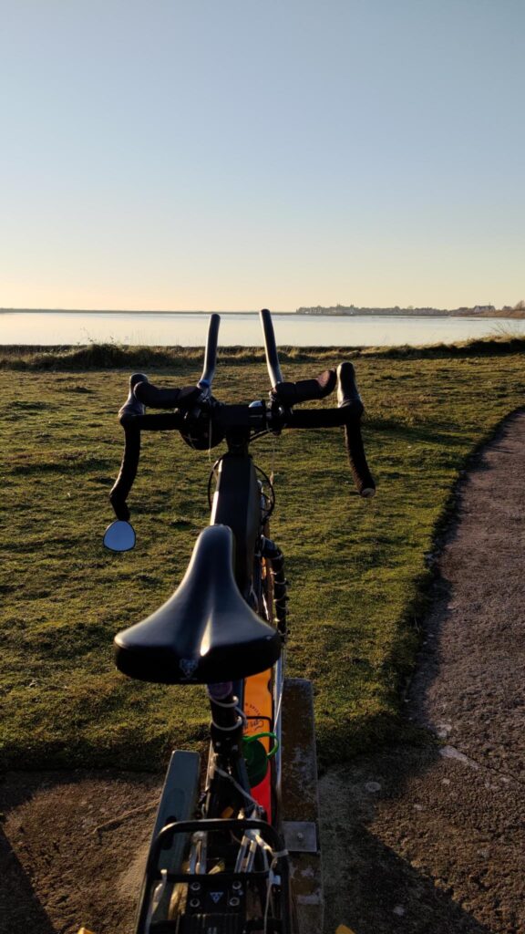Photo verticale, prise avec un vélo au premier plan et dans l'axe du vélo un plan d'eau et derrière le plan d'eau un village.