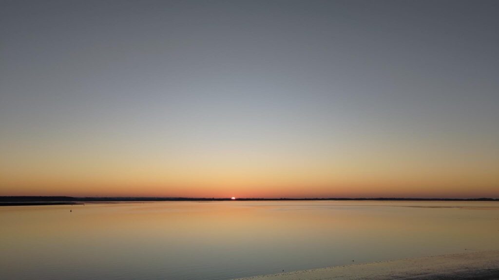 Photo d'un couché de soleil. On distingue au centre de l'image un petit point, c'est un demi soleil. Au premier plan la mer fait un miroir sans mouvement. L'horizon fait un trait sombre au tiers bas de l'image. Le plan d'eau reflète les couleurs du ciel, orangé, jaune, bleu, gris.
