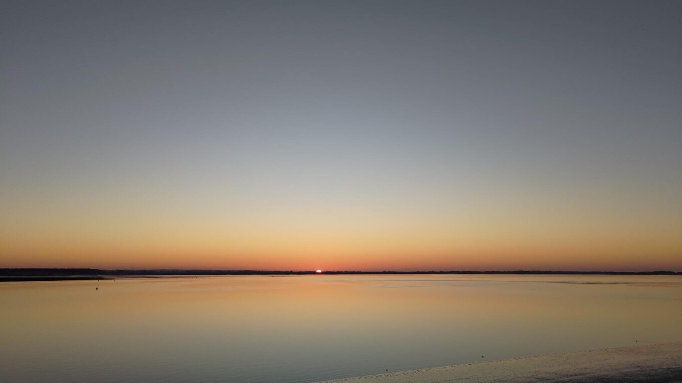 Photo d'un couché de soleil. On distingue au centre de l'image un petit point, c'est un demi soleil. Au premier plan la mer fait un miroir sans mouvement. L'horizon fait un trait sombre au tiers bas de l'image. Le plan d'eau reflète les couleurs du ciel, orangé, jaune, bleu, gris.