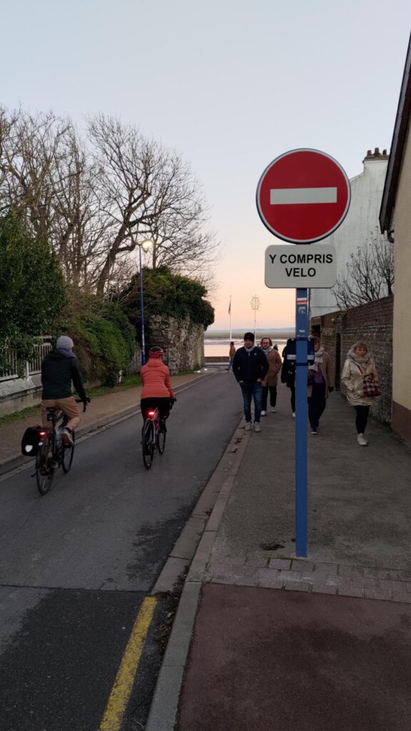 Une entrée de rue avec un panneau « sens interdit » et un petit panneau « y compris velo », deux cyclistes s'engagent dans cette rue