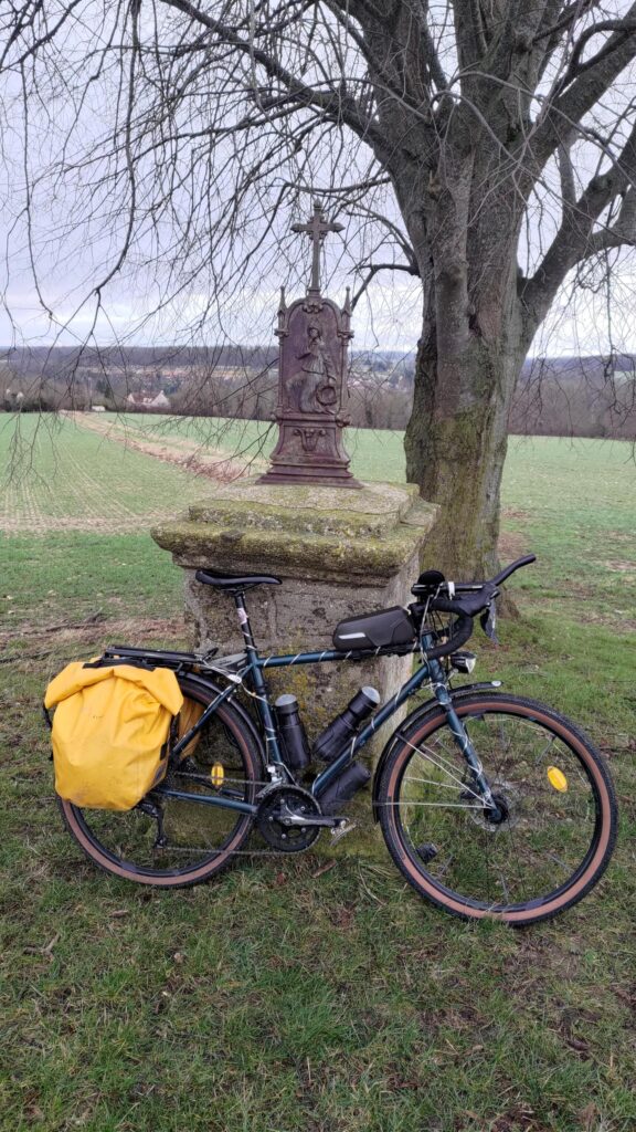 Un vélo type gravel est posé sur un socle de pierre portant une sculpture métallique religieuse représentant un personnage à moitié à genoux se masquant le visage avec la main gauche et tenant une couronne végétale dans sa main droite