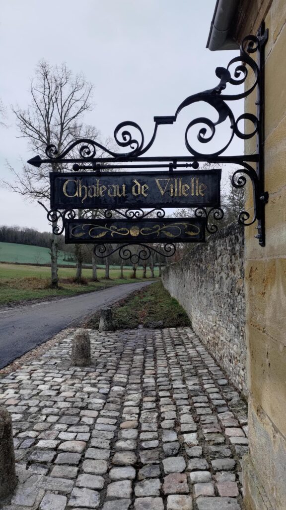 Enseigne forgée en métal indiquant en lettres dorées le Château de Villette, fixée au mur d'enceinte à l'entrée du château. Le sol est pavé. une petite rouge longe l'enceinte sur la gauche et un alignement d'arbres borde l'autre côté de la route.