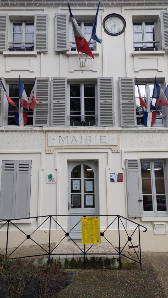 Plan serré de la façade de la mairie d'Évecquemont (78), petite bâtisse bourgeoise de deux étages avec des drapeaux français et européens sur plusieurs fenêtre. Un baromètre géant orne la façade entre deux fenêtres du deuxième étage.