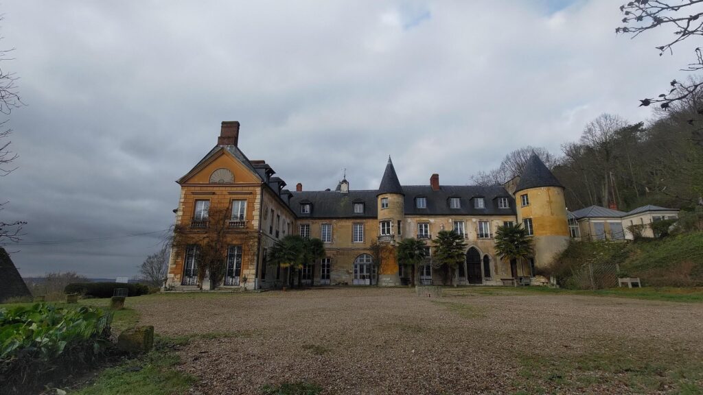 Cours en cailloux du château de Vaux sur Seine. C'est une grande bâtisse de couleur ocre et un toit en tuile, en forme de L. Deux étages et un étage sous comble. une petite tour avec un toit en cône est proéminente au centre de la façade, une seconde 2 fois plus large sur le côté droit. À gauche du bâtiment la vue est très dégagée car nous sommes à flan de coteaux. À droite le terrain monte en pente, une annexe est collée au château et on distingue une partie d'une grande serre. Des palmiers sont plantées dans la cours le long de la façade.